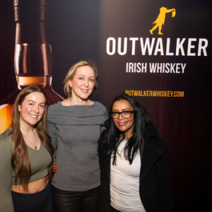 Three women posing for a photo next to an Outwalker Whiskey stand.