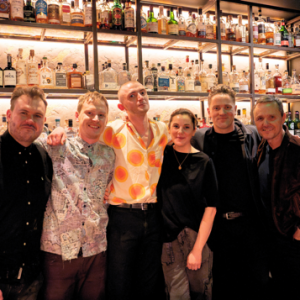 A group of people, five men and a woman, center, smiling in a row at a bar.